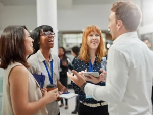 The image shows a group of four professionals engaged in a lively conversation. Three women and one man are standing in a modern, bright, open space, likely at a conference or networking event. They are all smiling and appear to be enjoying the discussion. The women are holding items like a tablet, notebook, and coffee cup, while the man holds a water bottle. The setting suggests a positive and collaborative environment.